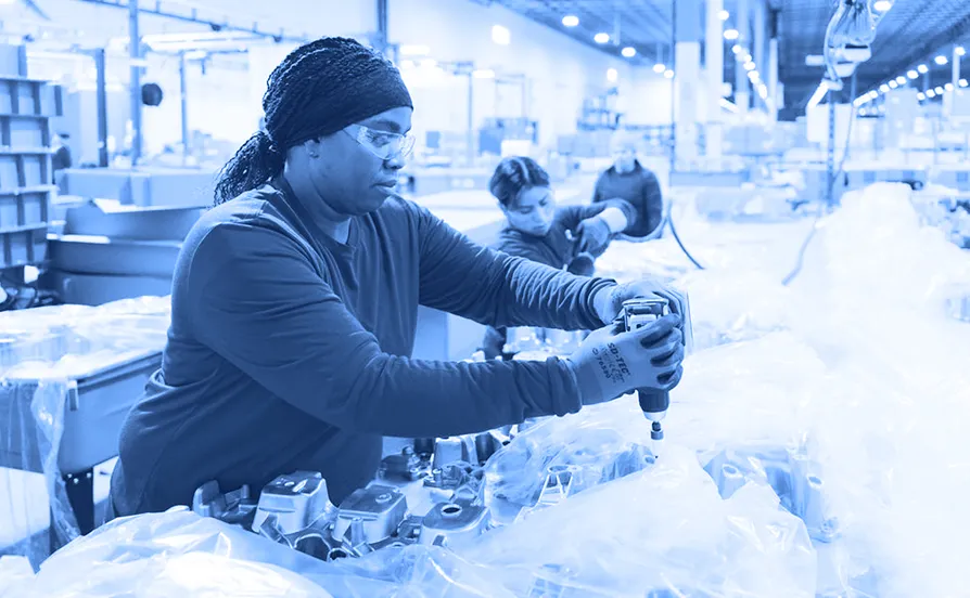 Worker wearing gloves and safety glasses using a handheld power tool to fasten components on plastic-wrapped metal parts at an assembly station inside a warehouse.