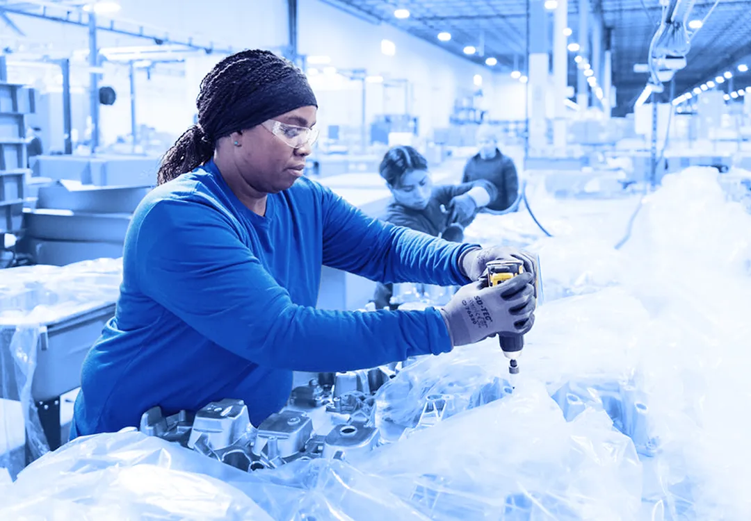 Worker wearing gloves and safety glasses using a handheld power tool to fasten components on plastic-wrapped metal parts at an assembly station inside a warehouse.