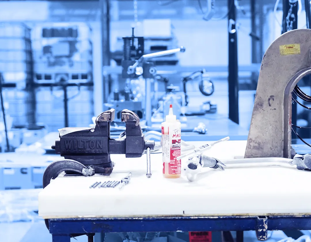 Workbench with a metal vise, hand tools, fasteners, and a bottle of adhesive arranged on a tabletop inside a factory area.