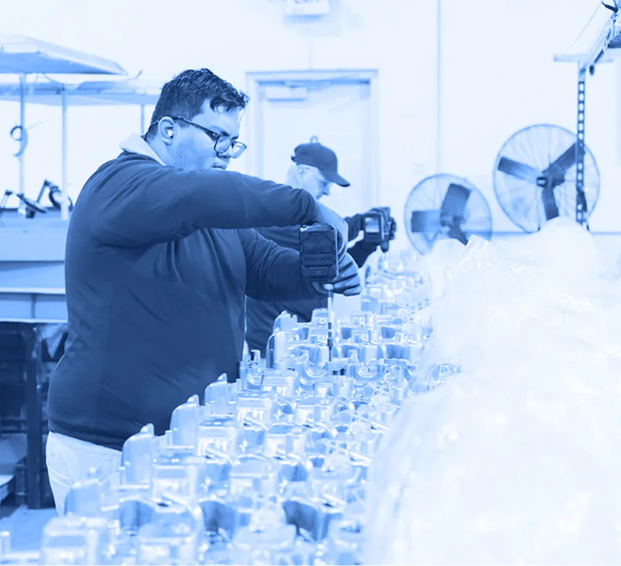 Worker in a blue shirt using a handheld power driver to fasten components on a row of metal parts at an assembly workstation, with packaging materials and equipment in the background.