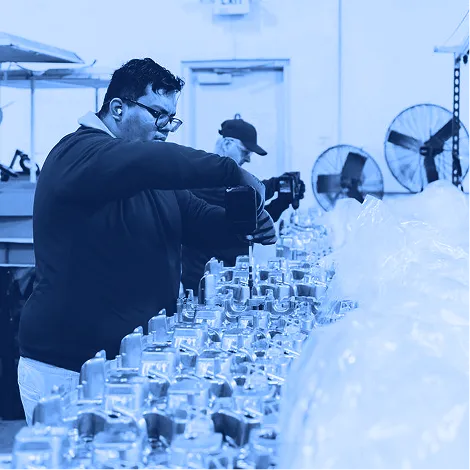 Worker in a blue shirt using a handheld power driver to fasten components on a row of metal parts at an assembly workstation, with packaging materials and equipment in the background.