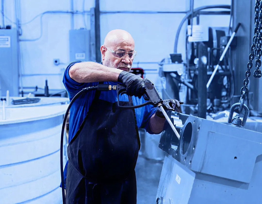 Worker wearing gloves and safety glasses using a handheld washing tool on a large metal component suspended by chains inside a factory workspace.