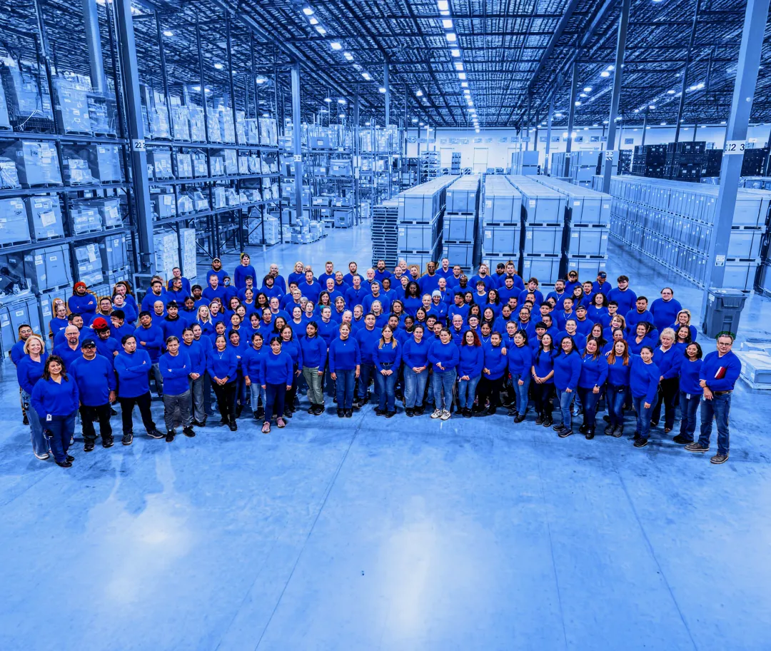 Large group of workers wearing blue shirts standing together on a warehouse floor with pallet racking and stacked boxes in the background.