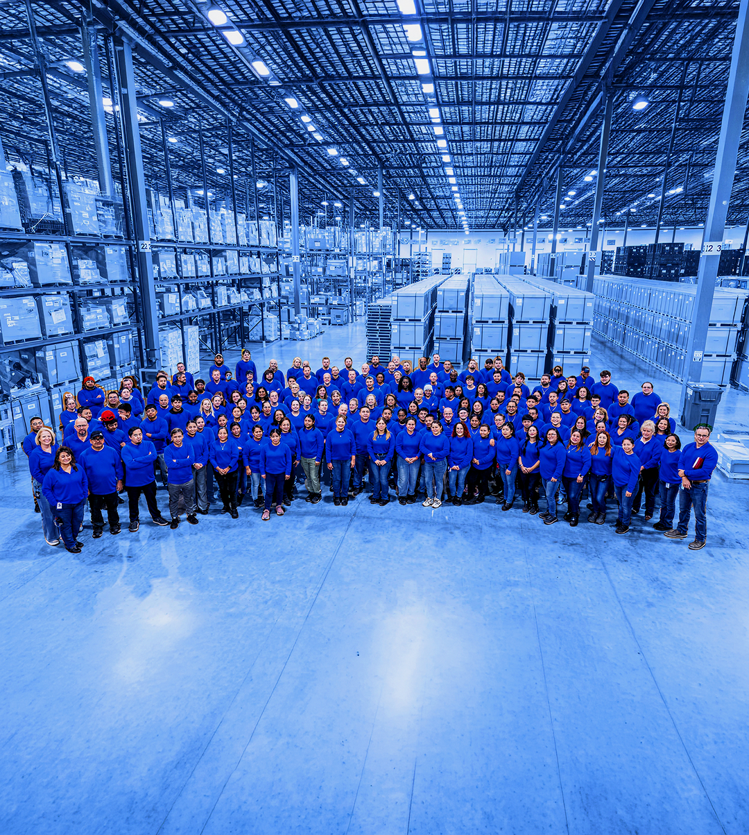 Large group of workers wearing blue shirts standing together on a warehouse floor with pallet racking and stacked boxes in the background.