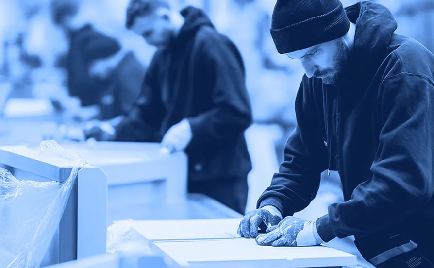 Workers wearing dark hooded sweatshirts and gloves assemble flat cardboard components at workstations inside a warehouse, with boxes and packing materials on tables in the foreground.