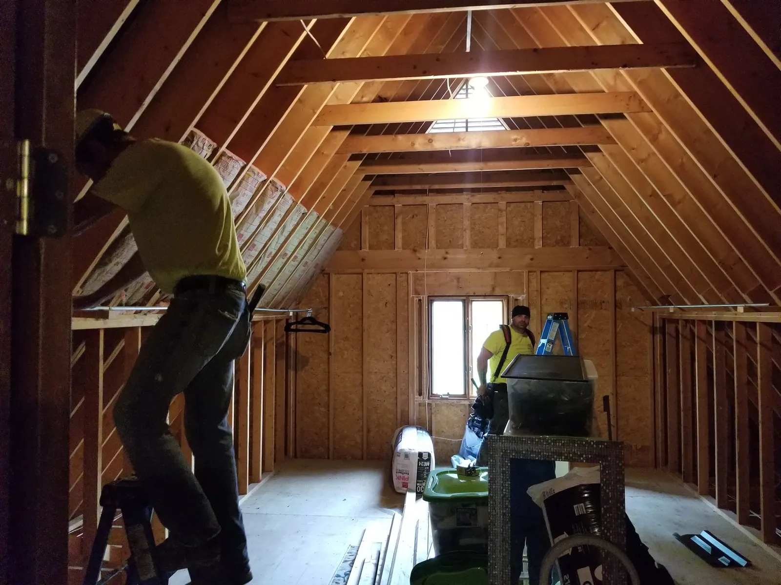 Two workers installing insulation along rafters in an unfinished attic with wooden framing.