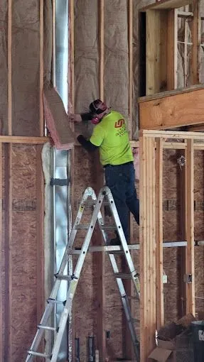 Worker in a neon green shirt installing fiberglass insulation inside a wooden frame house using a ladder.