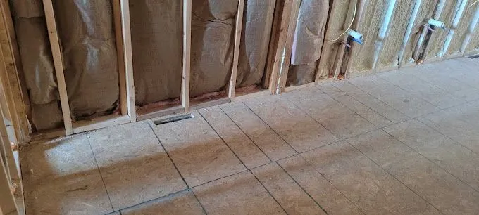 Interior wall framing with fiberglass insulation and unfinished subfloor marked with layout lines.
