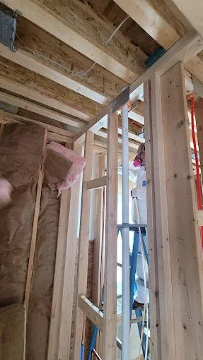 Construction worker in protective gear installing fiberglass insulation inside wooden frame walls of a building.