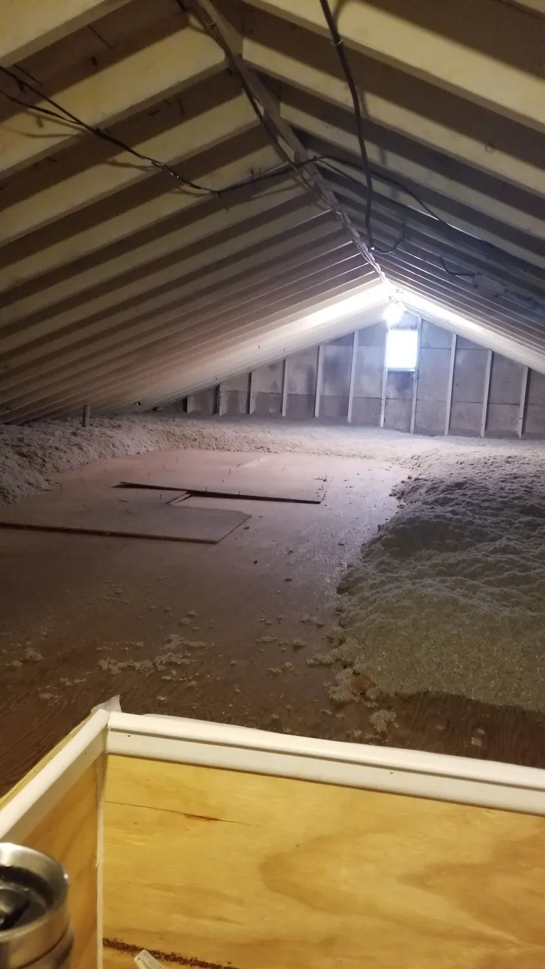 Attic interior with visible wooden beams and loose-fill insulation spread unevenly on the attic floor.