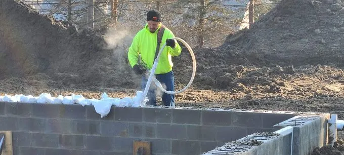 Worker spraying foam insulation on concrete blocks at a construction site outdoors.
