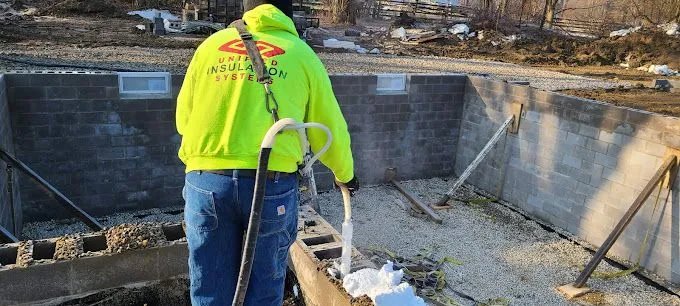 Worker in bright yellow-green hoodie pouring foam insulation from a hose into concrete blocks at a construction site.