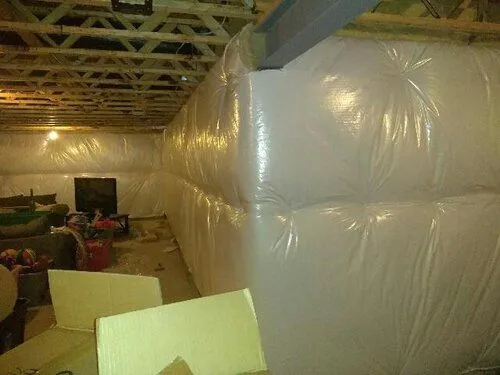 Basement with full wall and ceiling covered in protective white plastic sheeting wrapped around insulation or framing.