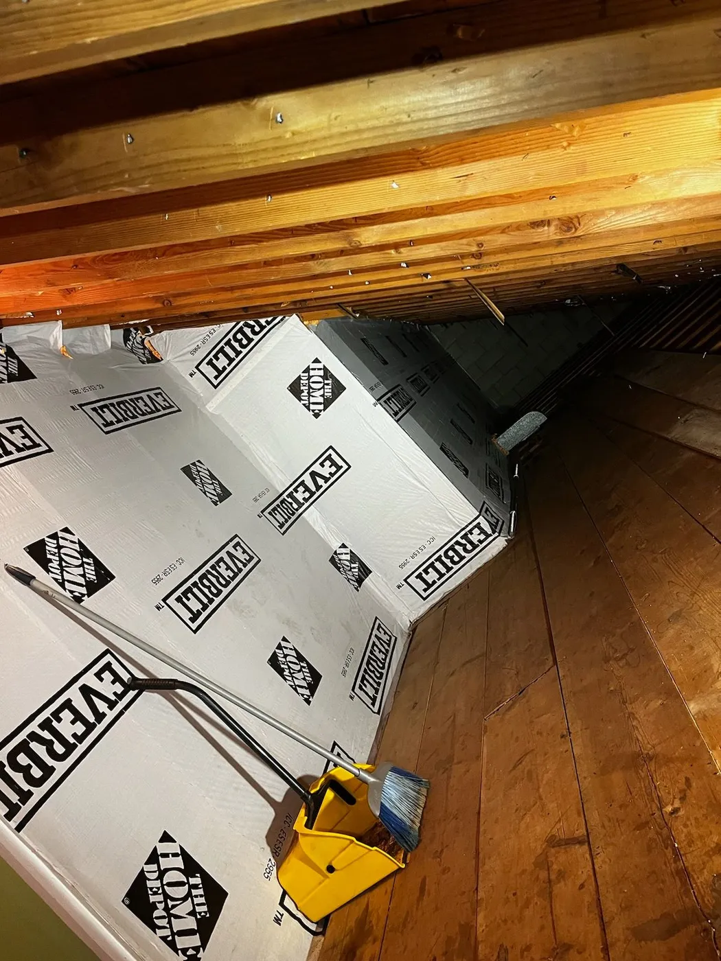 Attic space with wooden floor and beams, insulated walls covered in The Home Depot Everbilt insulation panels, and a yellow dustpan with a broom leaning against the wall.