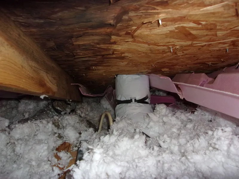 Old attic space with deteriorating white insulation scattered around ductwork under a wooden ceiling.