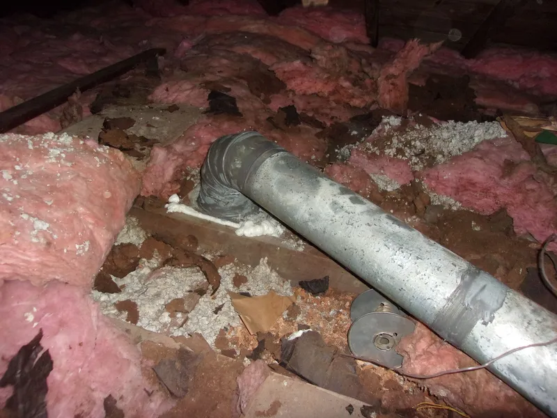 Damaged pink fiberglass insulation scattered around a metal duct pipe in an attic space.