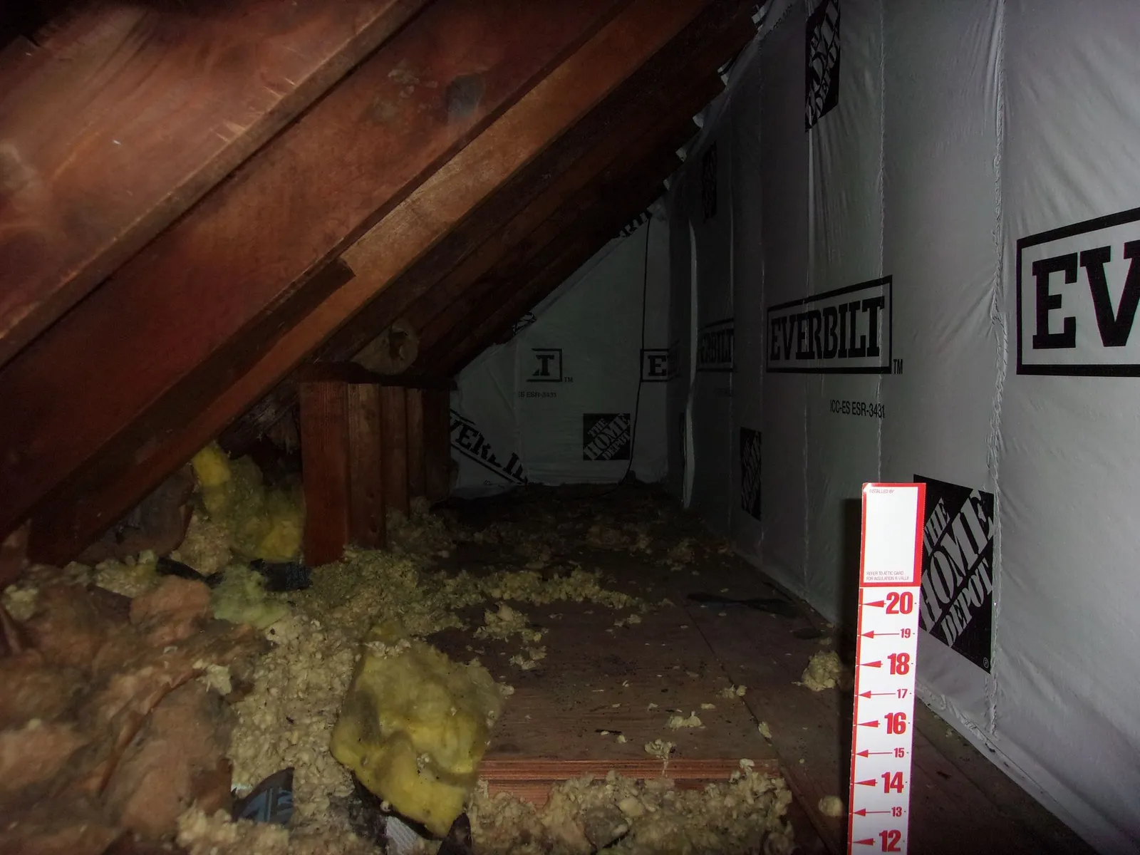 Attic space with exposed wooden beams and vapor barrier, scattered fiberglass insulation on floor with a measuring stick marked up to 20 inches.