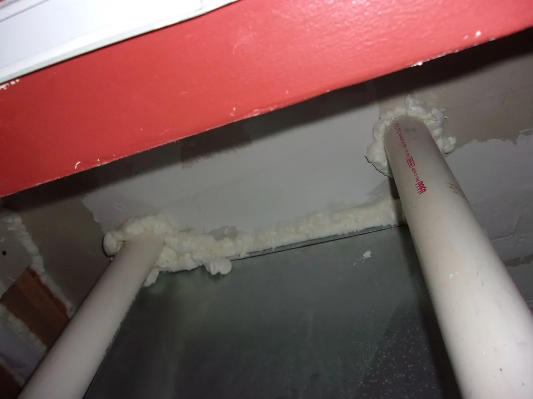Two white pipes penetrate a ceiling, sealed around the edges with white expanding foam insulation.