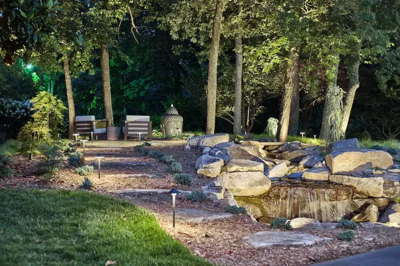 A beautifully lit garden path with stone steps, surrounded by lush greenery and trees. The pathway leads to a cozy seating area with chairs and a decorative water feature, creating a serene and inviting outdoor space.