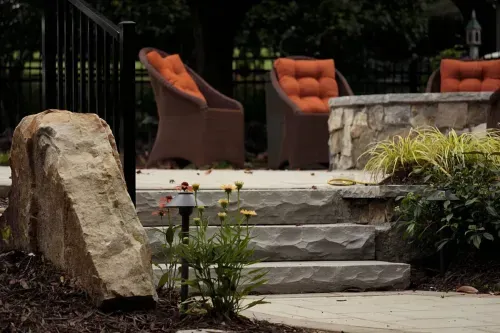 A close-up view of a hardscape design featuring large stone steps leading up to a patio area. The patio is adorned with comfortable wicker chairs with orange cushions, and a stone fire pit, offering a perfect spot for relaxation and gatherings.