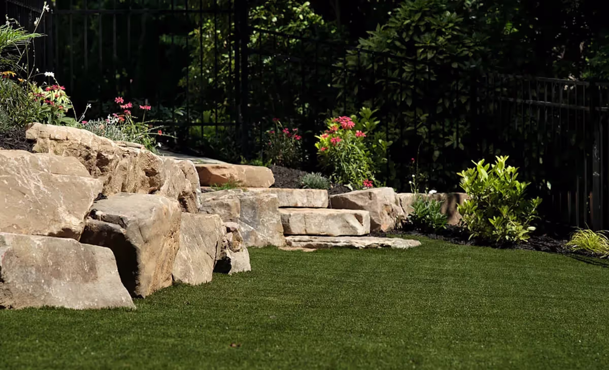 A landscaped garden with a series of stone steps and rock formations integrated into the grassy lawn. Colorful flowers and plants are strategically placed around the rocks, enhancing the natural beauty of the small garden space.