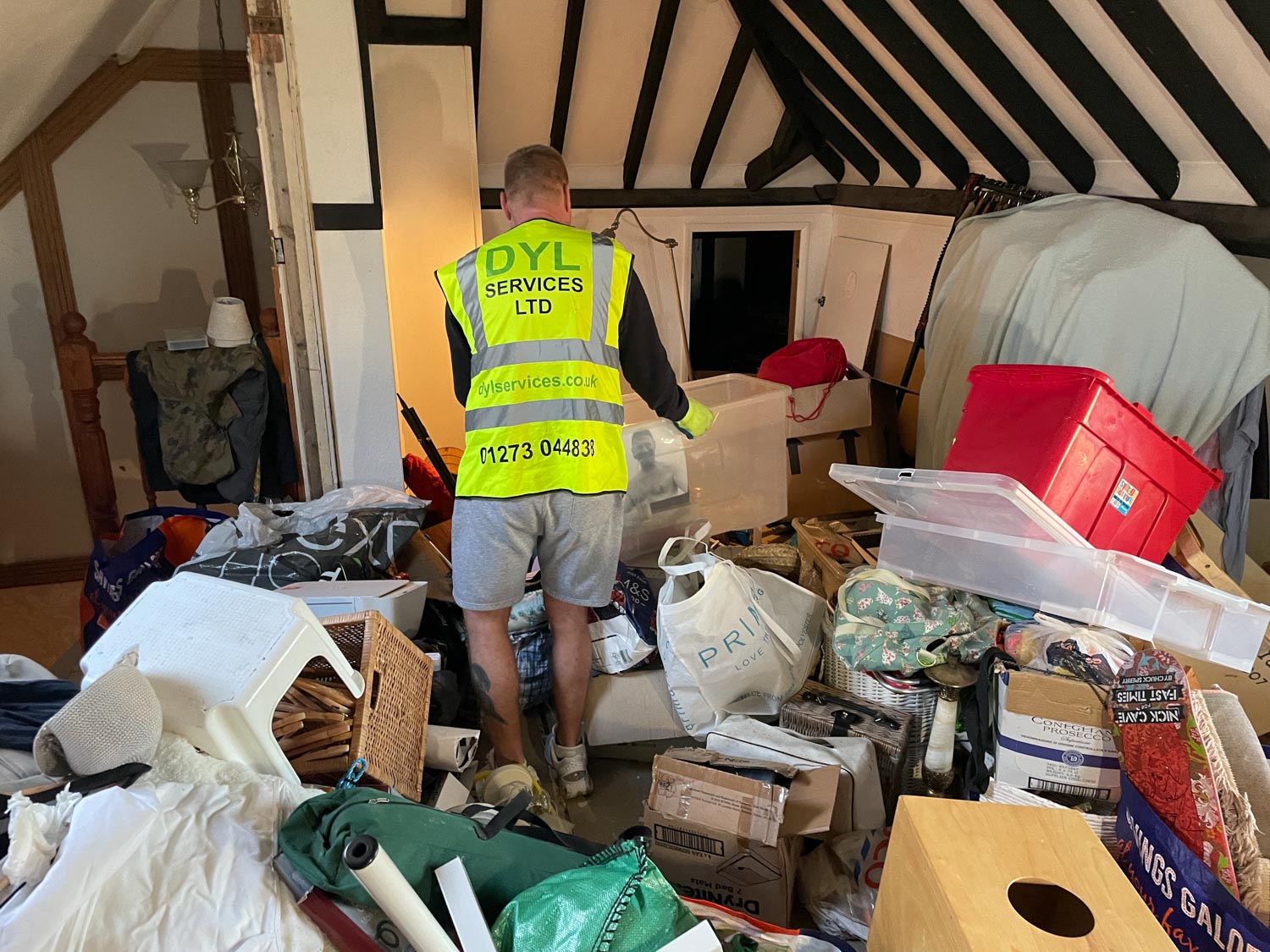 A worker in a high-visibility vest organizes a cluttered room filled with boxes and bags.