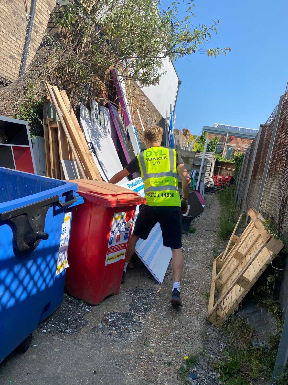 A person wearing a safety vest is moving a red bin in a narrow alleyway.
