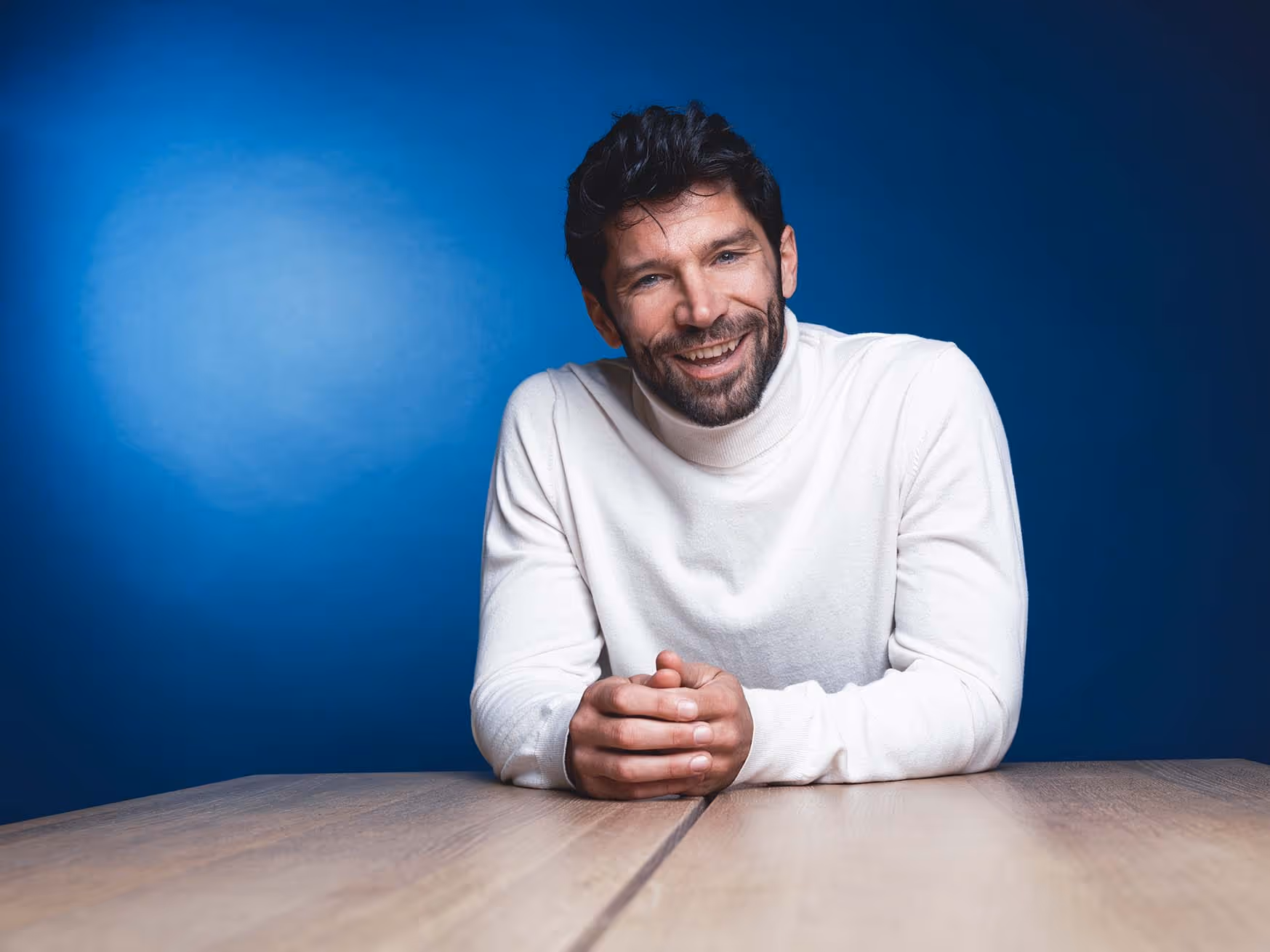 Lächelnder Mann mit dunkelbraunem Haar und Bart in weißem Rollkragenpullover vor blauem Hintergrund, der an einem Holztisch lehnt.