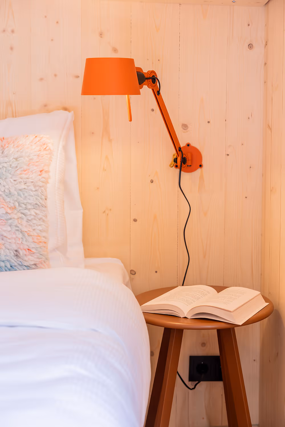 Oranje verstelbare leeslamp aan houten muur boven nachtkastje met open boek naast bed
