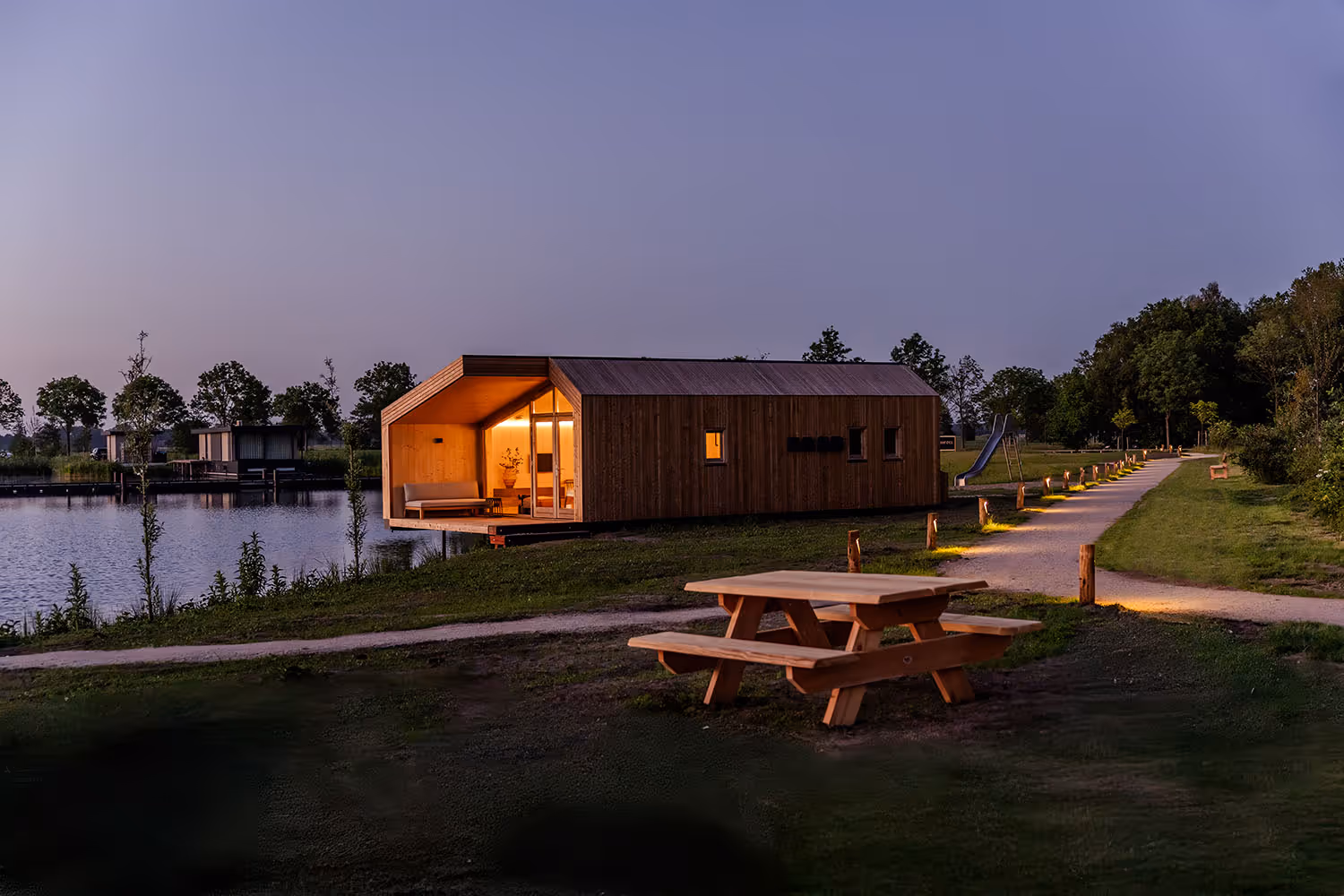Houten picknicktafel in grasveld met verlichte moderne houten huisje bij water in de avond.