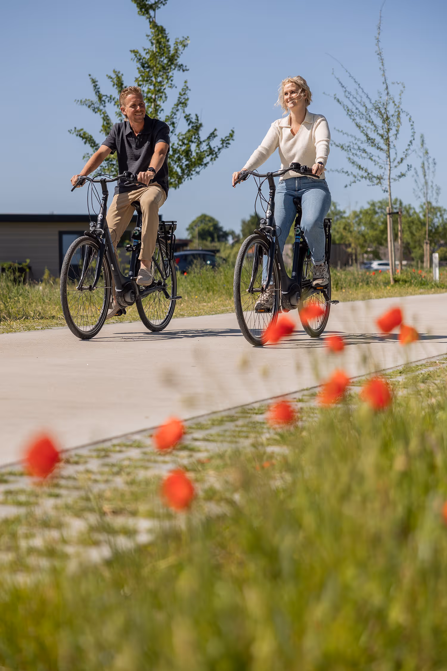 Man en vrouw fietsen op een zonnig pad met groene bomen en rode bloemen langs de weg. Ze zijn op vakantie bij vakantiepark Goudpoel in Friesland. 