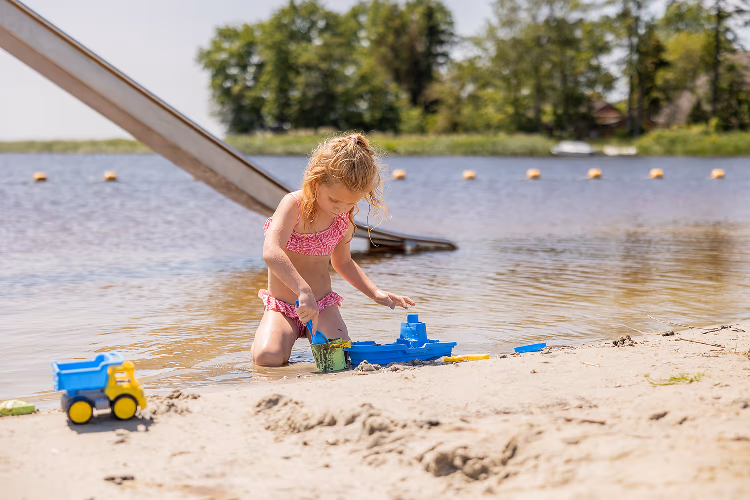 Meisje in roze bikini speelt met blauwe boot en zand op het strand bij het water bij vakantiepark Goudpoel.