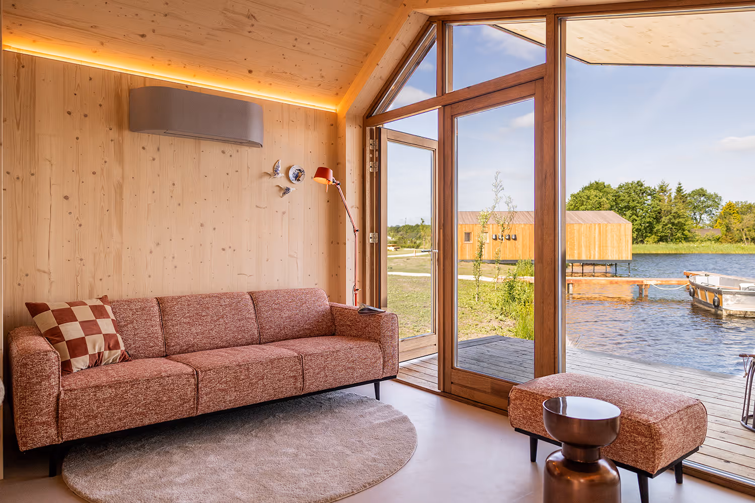 Gezellige zithoek met roze bank, poef, vloerlamp en uitzicht op een waterlodge aan het water bij Goudpoel in Friesland.