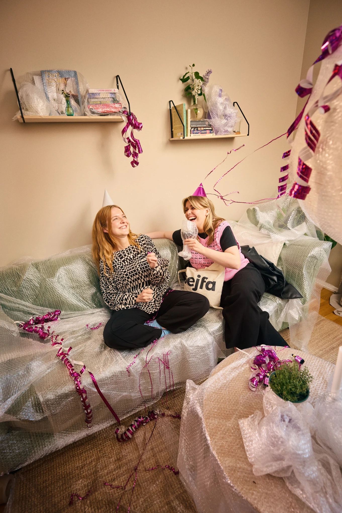 Två kvinnor skrattar under en fest i ett hem skyddat med bubbelplast – en lekfull symbol för trygghet och hemförsäkring från Sejfa.