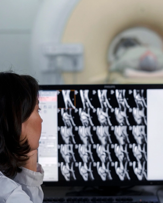 A medical professional in a white lab coat sits at a desk in a dimly lit control room, analyzing a grid of MRI scans on a computer monitor. Through a large observation window directly in front of them, a patient is visible lying on the bed of an MRI machine in the adjacent room.