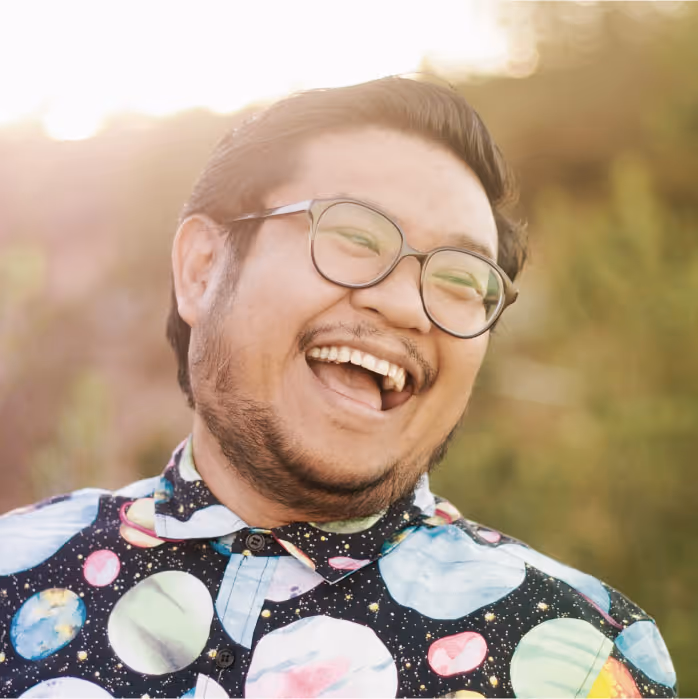 Man wearing glasses and a colorful shirt with planet designs laughing joyfully outdoors.