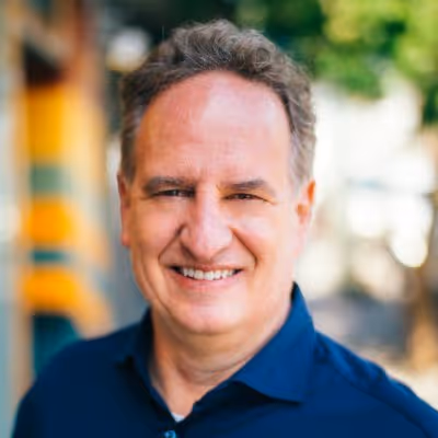 Smiling middle-aged man with short curly hair wearing a dark blue shirt, outdoors with blurred background.