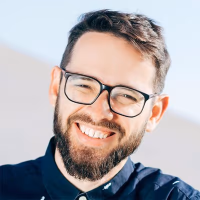 Smiling man with dark hair, beard, and glasses wearing a dark shirt against a light background.