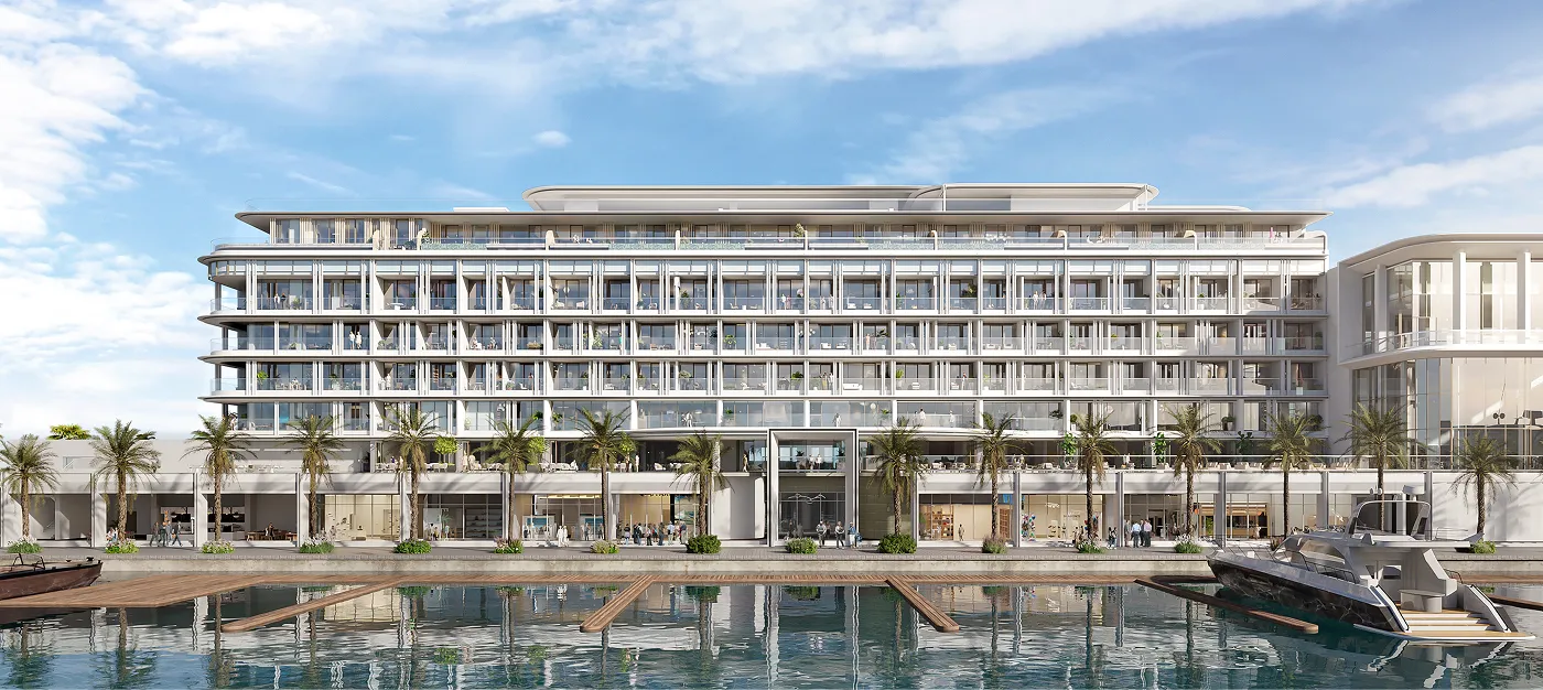 A modern hotel building featuring five floors with numerous balconies, situated along a waterfront. Palm trees line the front, and the structure's sleek design is complemented by a clear blue sky above.