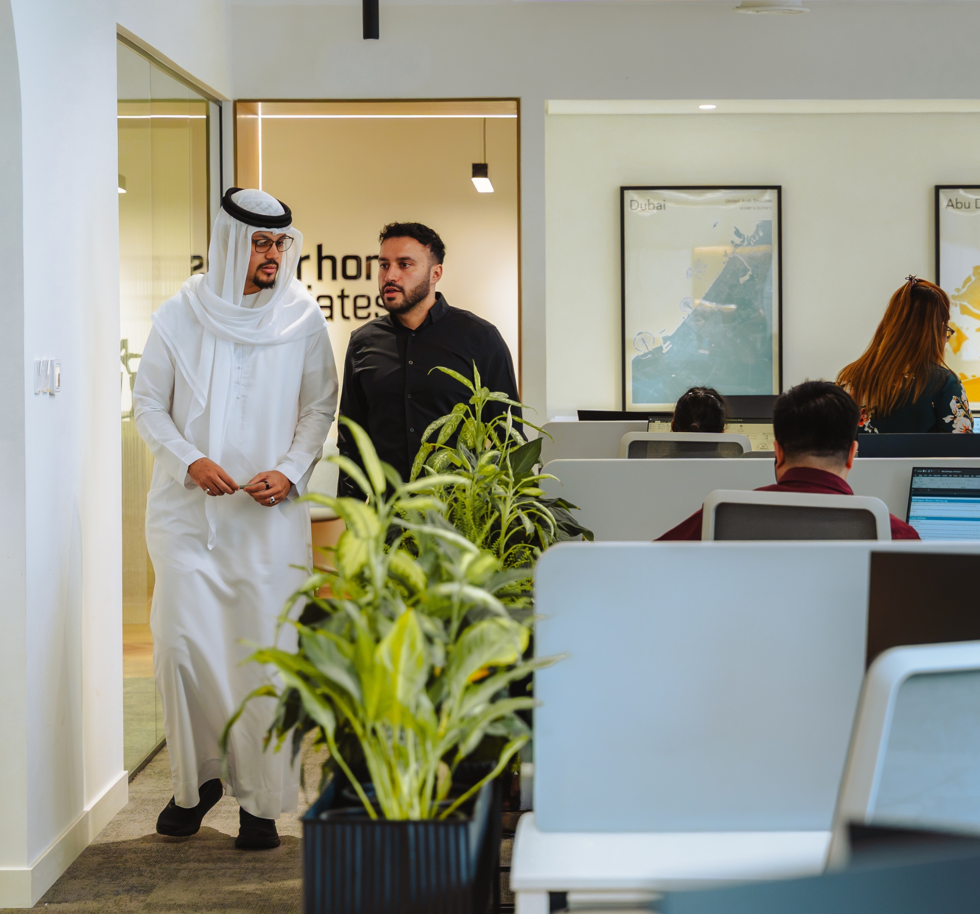 Man in traditional Middle Eastern white dishdasha visiting modern office with colleagues at workstations.