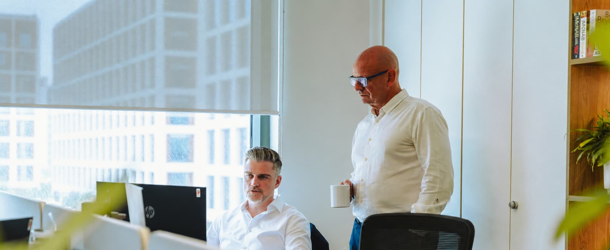 Two men in white shirts discussing work at a modern high-rise office desk.