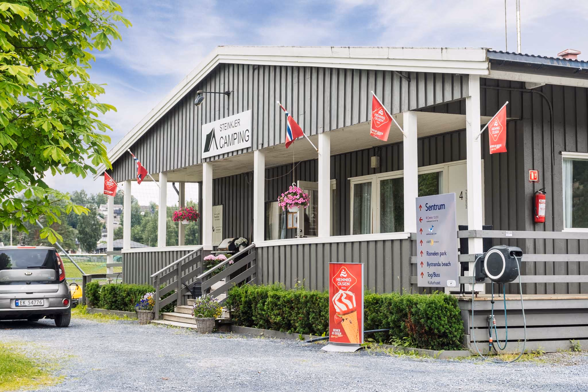 Grå bygg med Steinkjer Camping-skilt, blomster, isreklame og parkert bil utenfor.