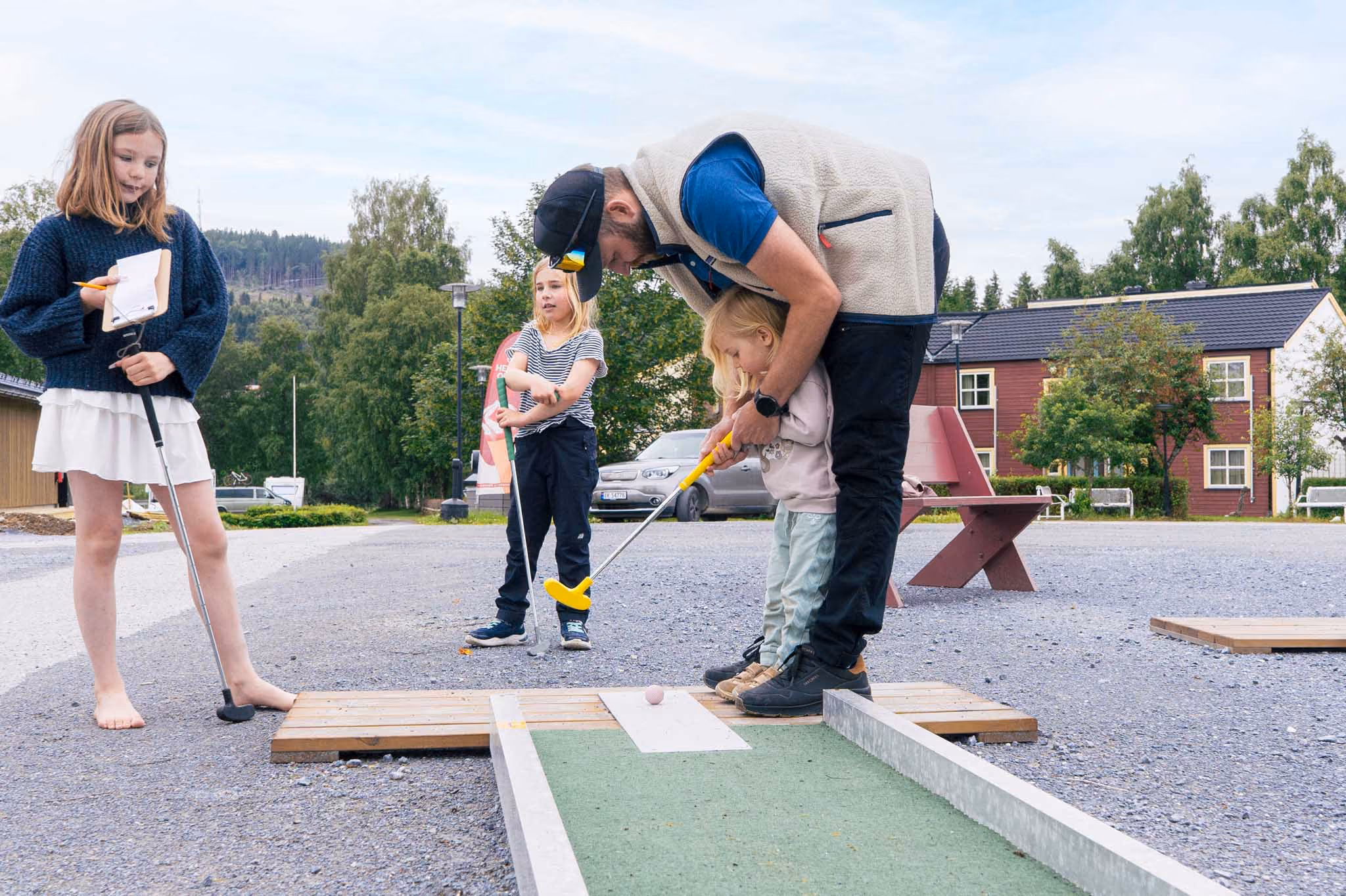En mann hjelper et lite barn med å spille minigolf mens to barn ser på utendørs.