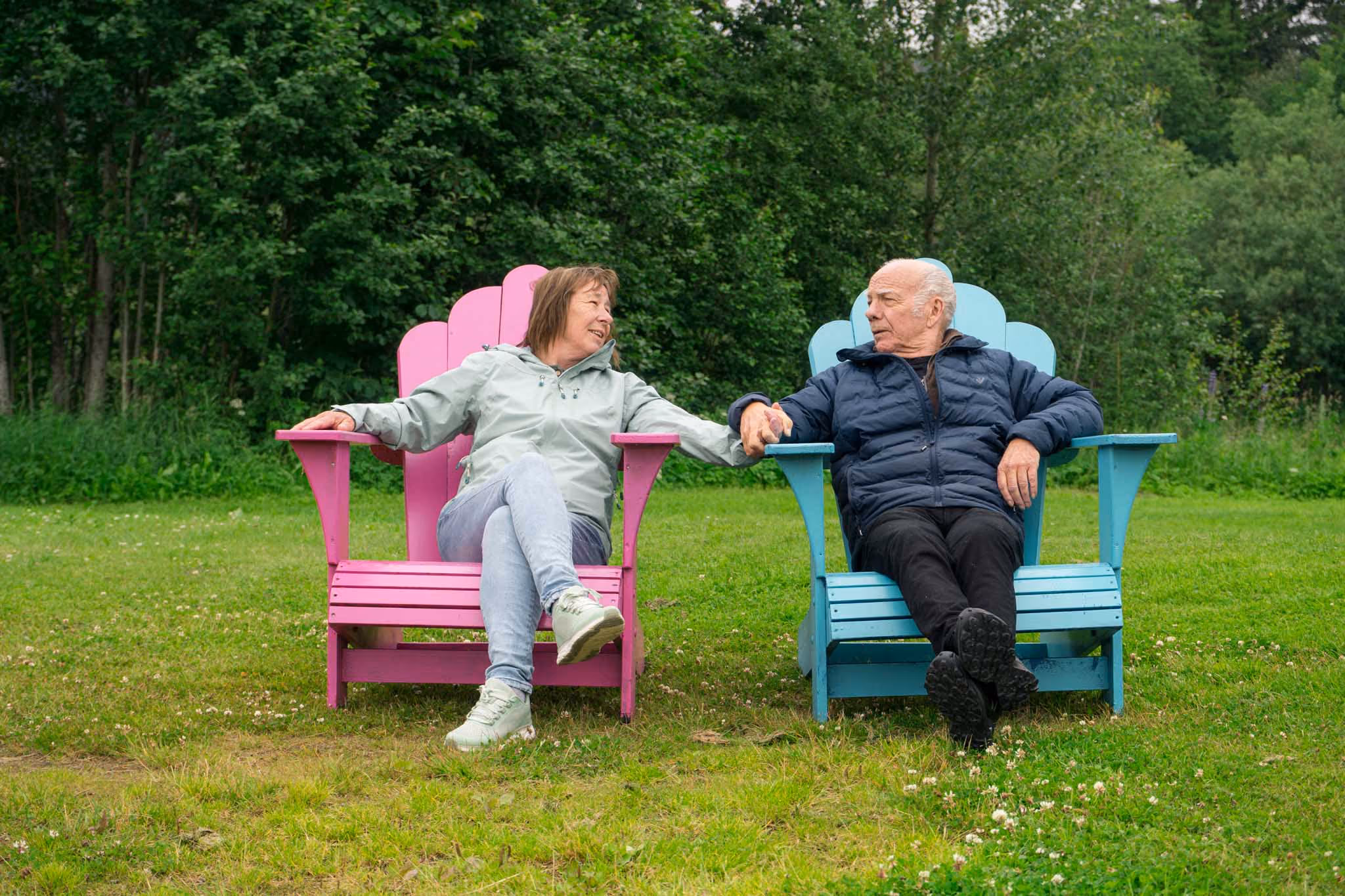 Eldre par sitter i rosa og blå hage-stoler, holder hender og ser på hverandre utendørs.