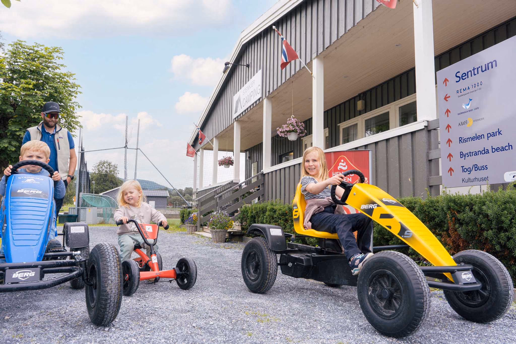 Barn kjører blå, gul og rød pedalbil utenfor Steinkjer Camping-bygning på en solrik dag.