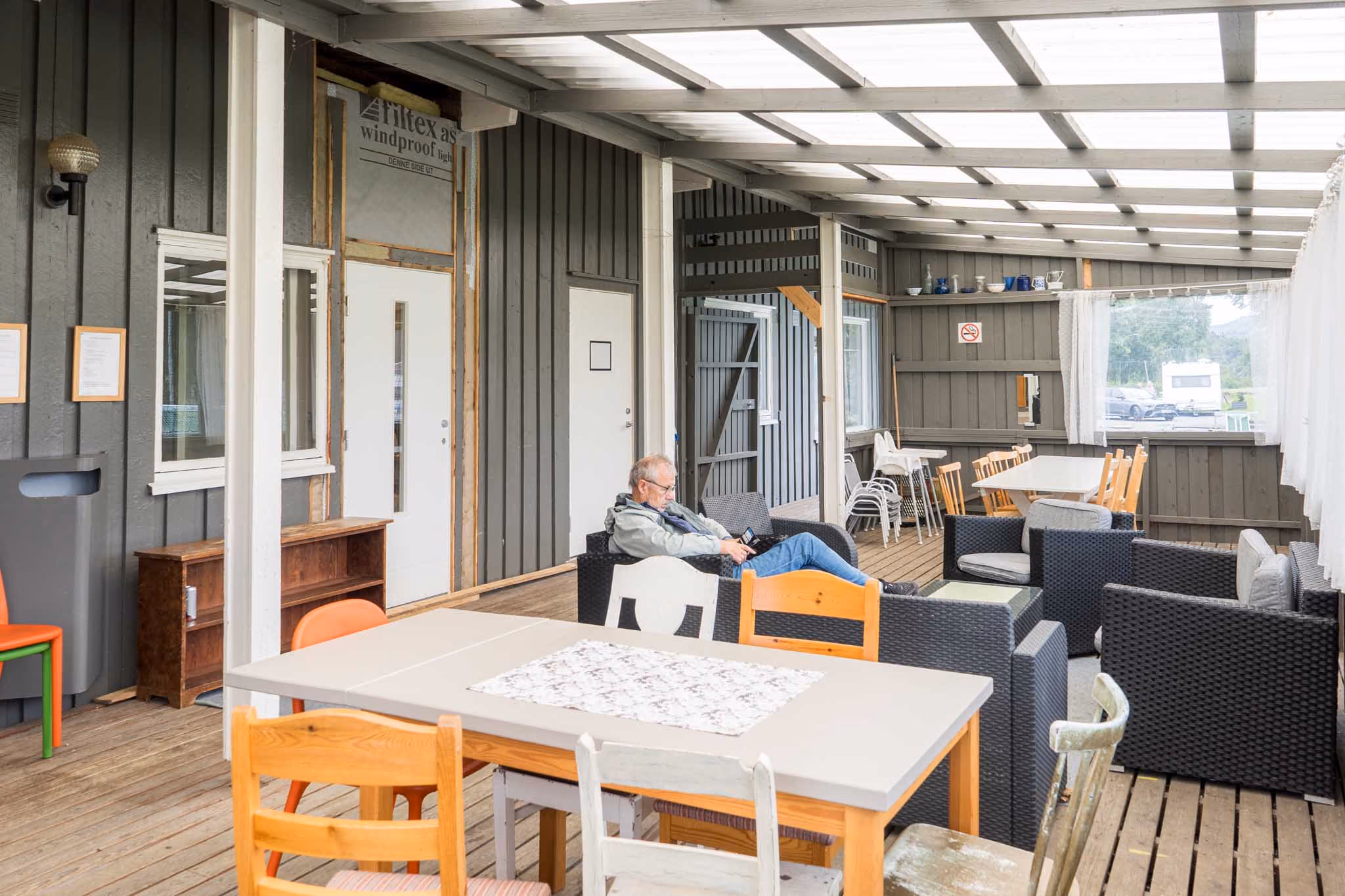 En eldre mann sitter alene på en stol i en innendørs terrasse med flere bord og stoler.
