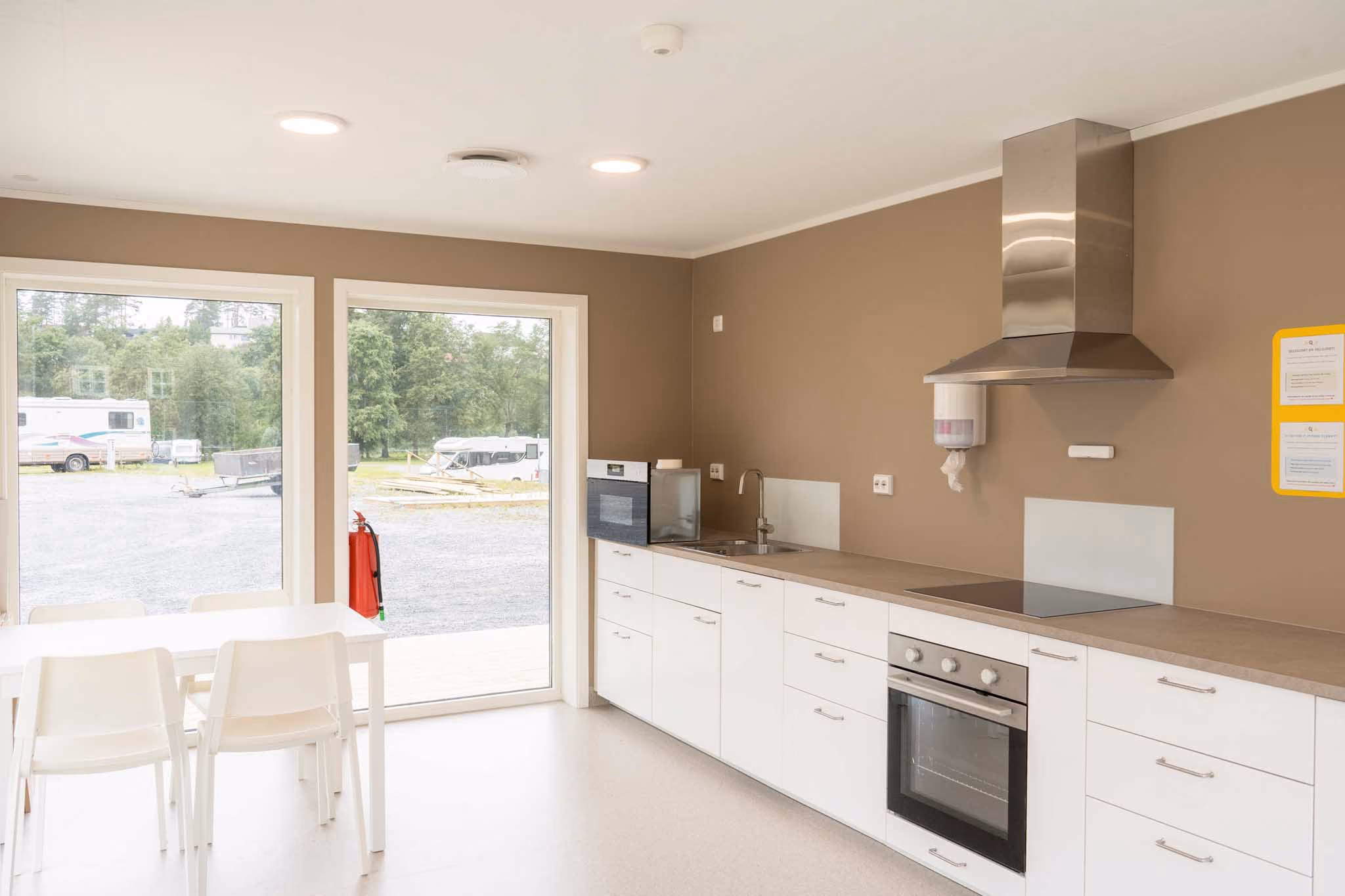 Lyst kjøkken med hvite skap, ovn, kokeplate, ventilator og spiseplass ved vindu med utsikt til camping.