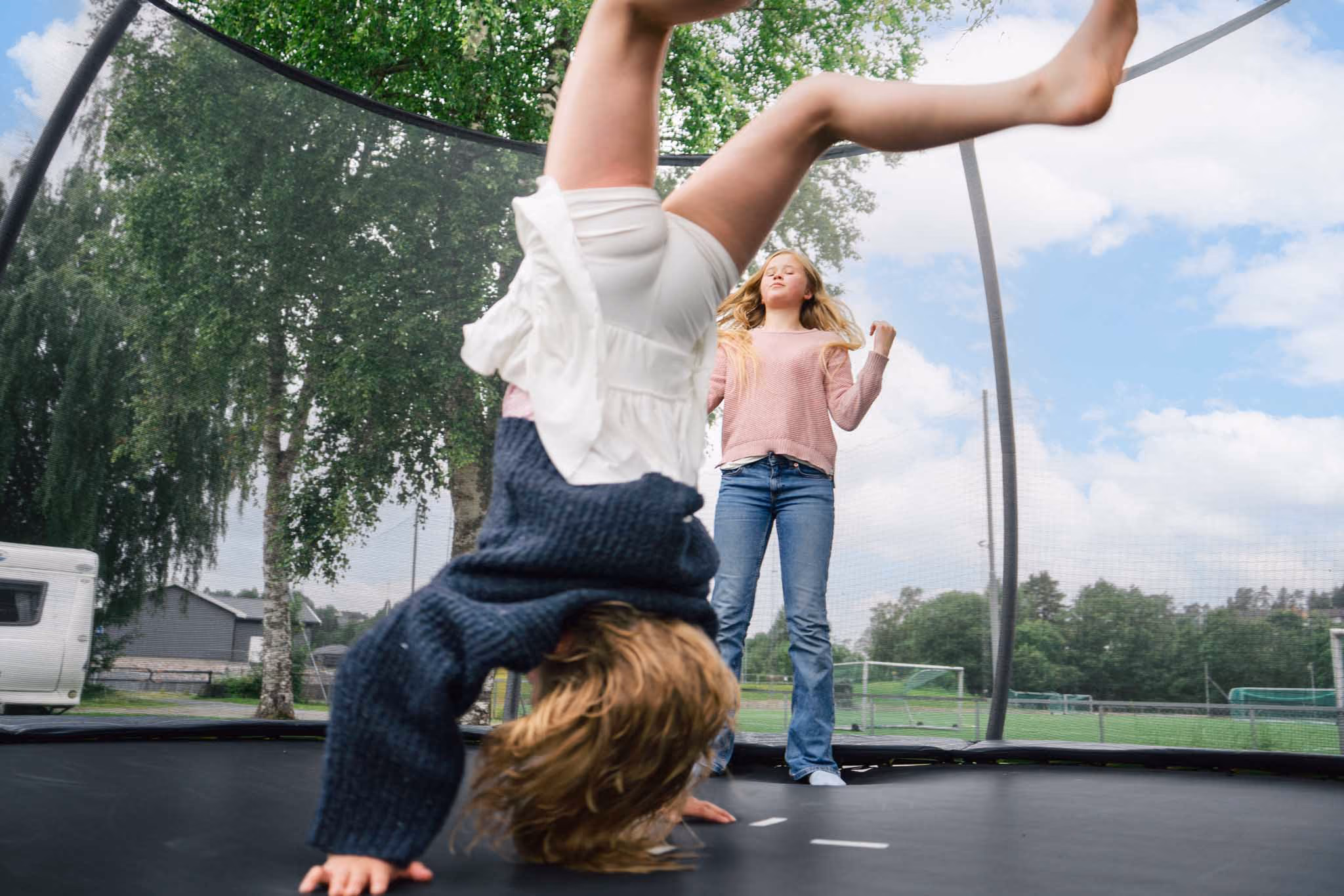 To jenter hopper og leker på en trampoline utendørs under blå himmel med trær i bakgrunnen.