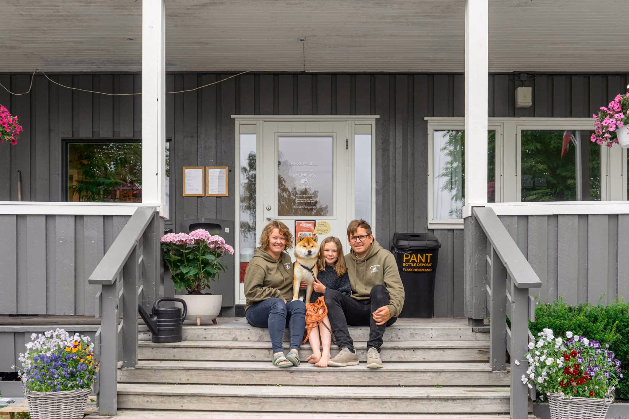 Familie med barn og hund sitter på trapp foran grå bygning med blomster og pantebøtte.