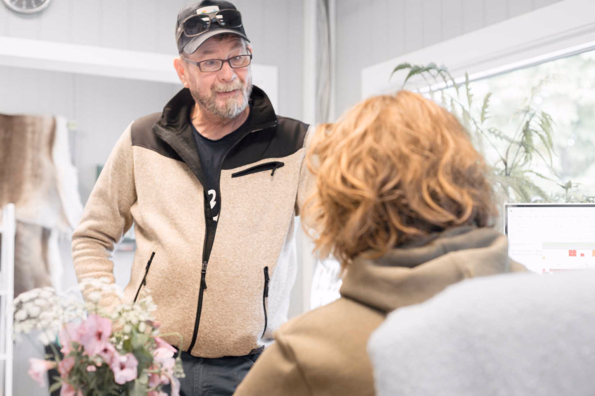 Mann med caps og briller snakker med en person med rødlig hår foran en datamaskin.